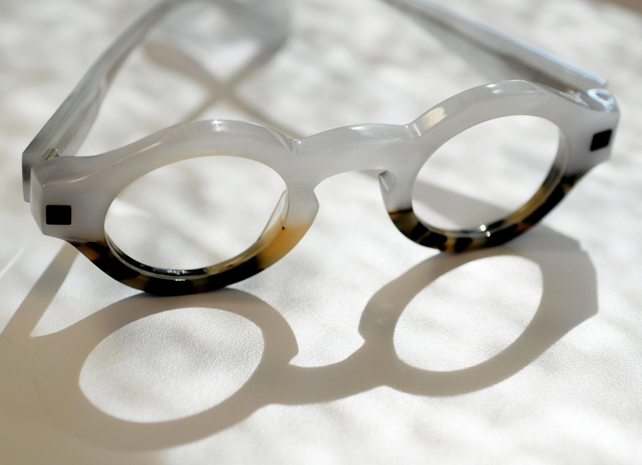 Editorial macro of a round pearl-white and tortoise acetate frame resting on white linen, casting a soft double-lens shadow.