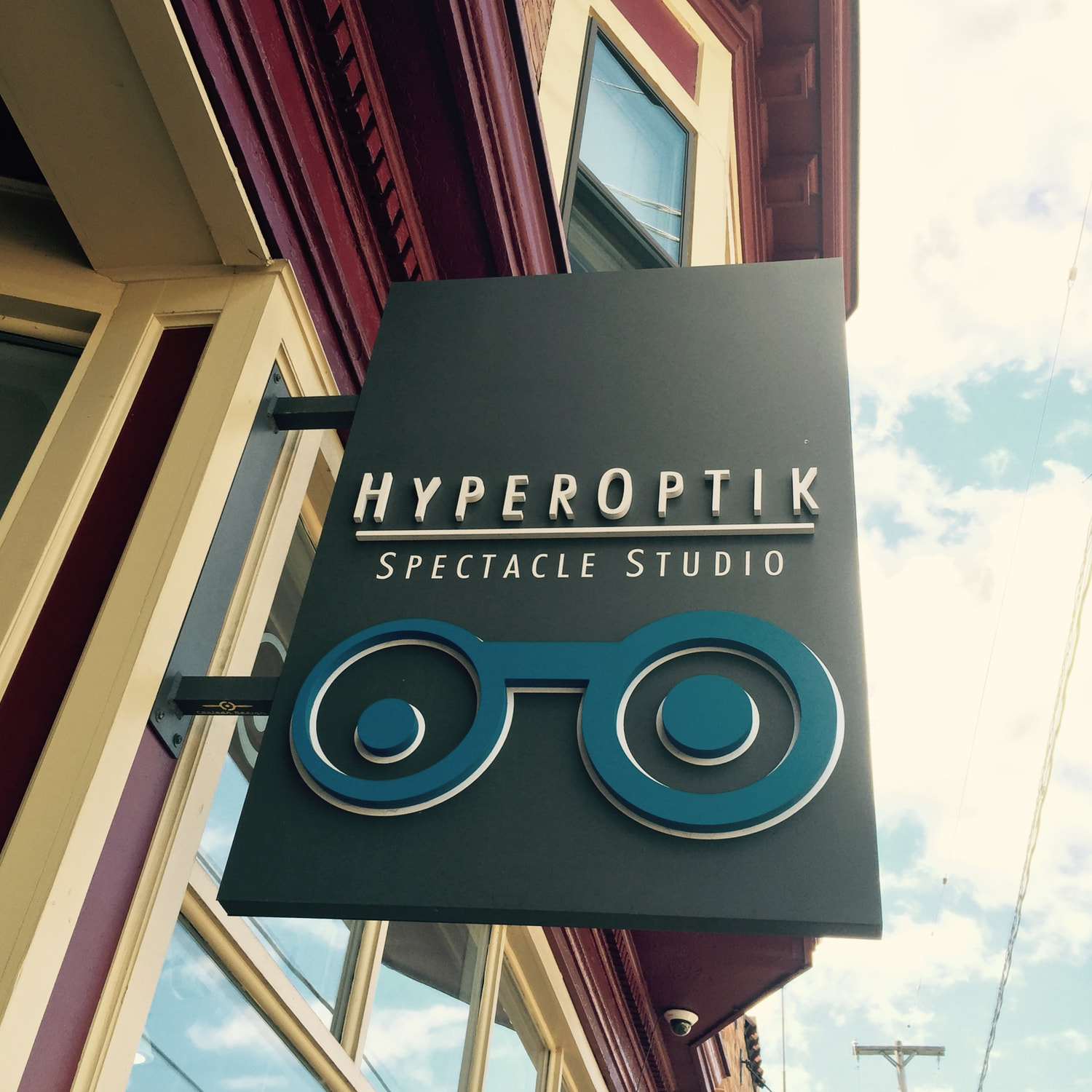 The HyperOptik Spectacle Studio blade sign mounted on the brick storefront against a bright sky.
